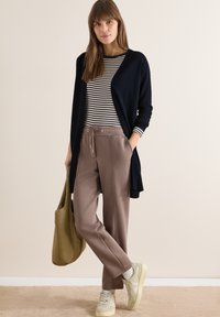 Femme debout en tenue décontractée avec une chemise rayée, un cardigan bleu marine, un pantalon taupe à cordon, des baskets beiges, et tenant un sac fourre-tout olive.