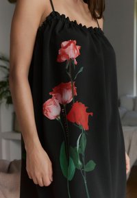 Zwarte jurk met een geplooide hals en dunne schouderbandjes, versierd met een bloemmotief van rode en roze rozen tegen een textuurachtergrond.