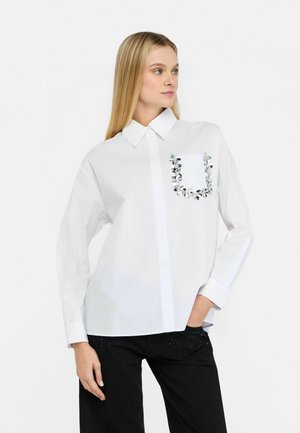 Camicia bianca con colletto, dotata di una tasca decorata con ornamenti d'argento. Maniche lunghe e taglio dritto.