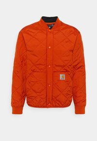 Rutig orange jacka med rund halsringning, tryckknappar, två framfickor och en logotyp på vänster ficka. Har ribbade ärmslut.