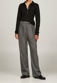 Schwarzes Polohemd mit langen Ärmeln, Knopfleiste und Logo, kombiniert mit grauen, plissierten Hosen aus strukturiertem Stoff und weitem Bein.