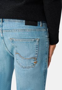 Jasnoniebieskie dżinsy mają tylną kieszeń z krzywym wzorem przeszycia, czarną skórzaną metkę przy pasie oraz prosty krój.