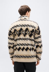 Μπεζ φλις πουλόβερ με μαύρα γεωμετρικά σχέδια, ψηλό γιακά και μακριά μανίκια. Απαλή υφή, άνετη εφαρμογή και διακοσμητικές ραφές.