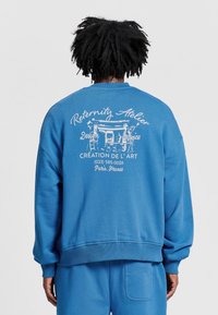 Niebieska bluza z okrągłym dekoltem, z prążkowanymi mankietami i dołem, z białym nadrukiem tekstowym i wzorem na plecach. Miękka tekstura materiału.