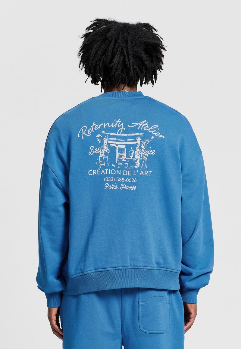 Niebieska bluza z okrągłym dekoltem, z prążkowanymi mankietami i dołem, z białym nadrukiem tekstowym i wzorem na plecach. Miękka tekstura materiału.