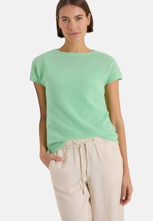 Femme portant un haut tricoté vert menthe à manches courtes et un pantalon beige à cordon, debout avec une main dans la poche, sur fond blanc.