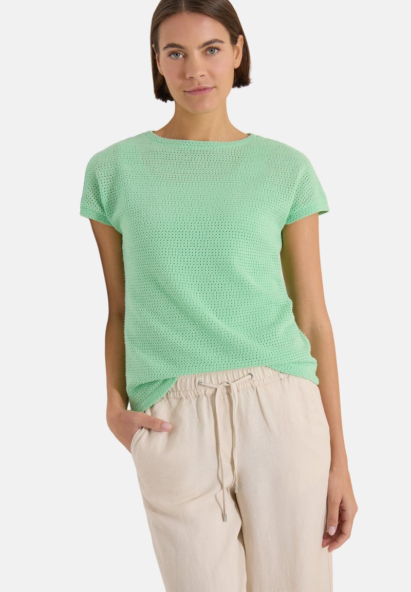 Femme portant un haut tricoté vert menthe à manches courtes et un pantalon beige à cordon, debout avec une main dans la poche, sur fond blanc.
