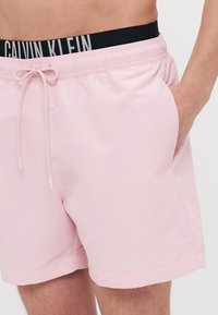 Helle rosa Badehosen aus glattem Stoff. Mit einem elastischen Bund, der ein schwarzes und graues "Calvin Klein"-Logo sowie einen Kordelzug aufweist.