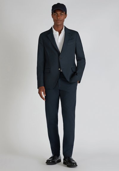 Marineblauem Anzug mit einem taillierten Blazer und passenden Hosen. Weißes Hemd mit Kragen darunter. Schwarze minimalistische Schuhe und eine schwarze Mütze.