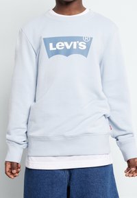 Personne portant un sweat-shirt Levi's bleu clair sur une chemise blanche et un jean bleu, les mains détendues sur les côtés contre un fond uniforme.