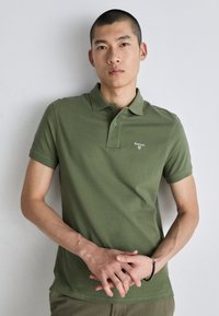 Olivazöld pólóing, pamutból készült, klasszikus gallérral, rövid ujjal és diszkrét logóval a bal mellen. Minimalista dizájn.