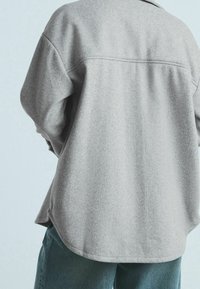 Graues, strukturiertes Stoffhemd mit lockerer Passform, abgerundetem Saum und Rückenpasse. Kombiniert mit dunkelgrünen Jeans, von hinten gezeigt.