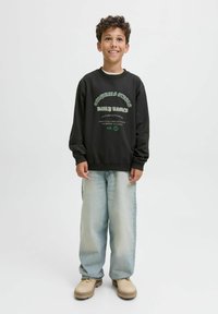 Felpa nera con design di testo bianco e verde, abbinata a jeans baggy blu chiaro e scarpe beige. Tessuto morbido, vestibilità casual.