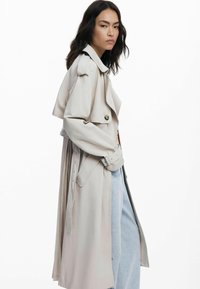 Light beige trench coat with structured shoulders, a double-breasted front, waist belt, and wide collar, paired with light blue jeans.