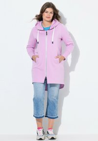 Sweat à capuche violet clair avec une fermeture éclair complète, cordons blancs, poches latérales, et un dos plus long, associé à un pantalon en denim court.