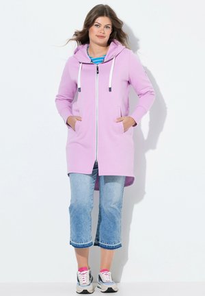 Felpa cappuccio di colore viola chiaro con zip completa, lacci bianchi, tasche laterali e un orlo posteriore più lungo, abbinata a pantaloni in denim cropped.