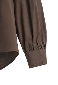 Camicia marrone a maniche lunghe con polsini arricciati e orlo leggermente curvato. Presenta la scritta "POLO CLUB" ricamata sul polsino. Tessuto morbido al tatto.