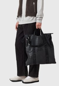 Bolso tote de cuero negro con branding en relieve, diseño grande, doble asa y acabado texturizado; llevado por una persona con atuendo casual.