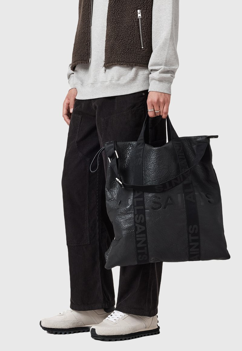 Bolso tote de cuero negro con branding en relieve, diseño grande, doble asa y acabado texturizado; llevado por una persona con atuendo casual.