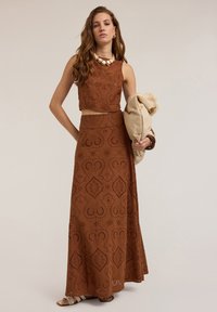 Raitapari ruskea kaksiosainen asu, jossa on hihaton yläosa ja pitkä hame, jossa on hienoja leikkuukuviota. Kankaan pinta on pehmeä ja muotoilu liehuva.