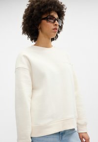 Krémová mikina s kulatým výstřihem, lehký materiál, žebrované manžety a lem, s diskrétním logem na hrudi.