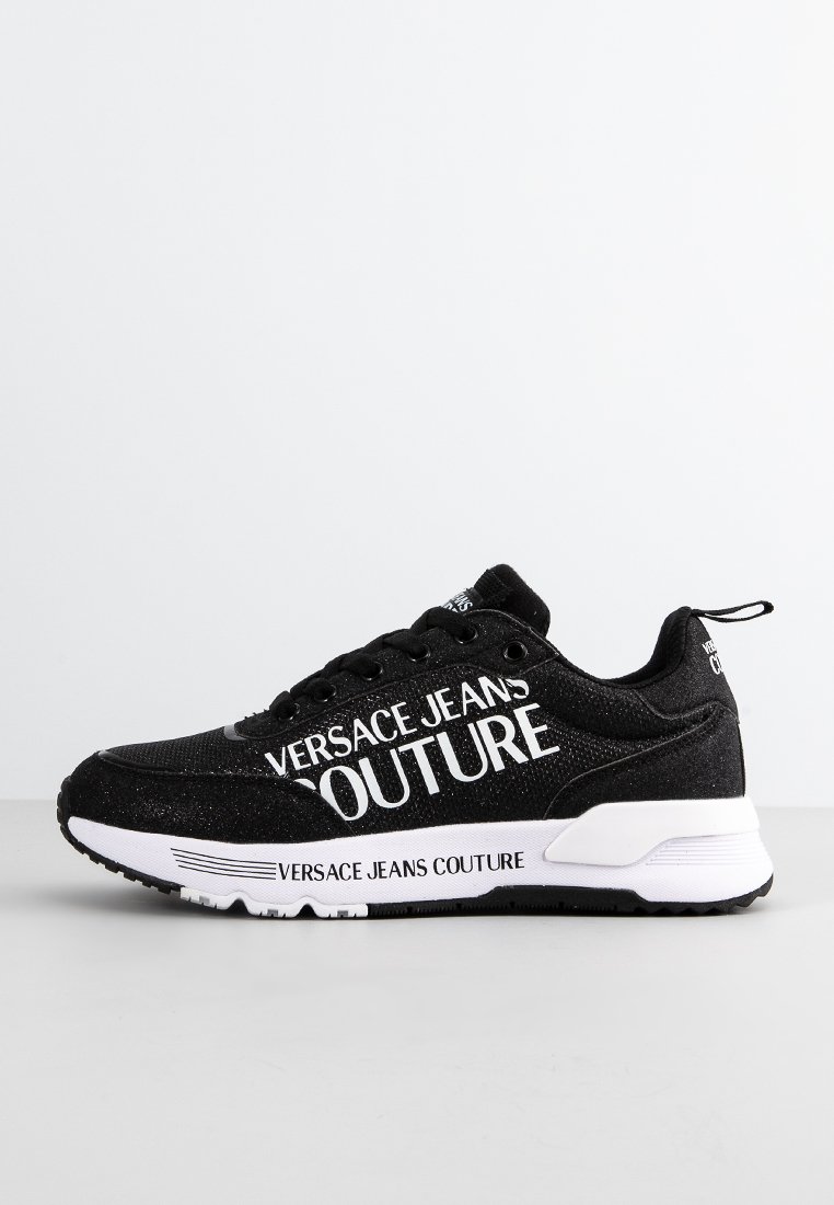 Zapatillas negras con la marca "VERSACE JEANS COUTURE" en blanco, tejido texturizado, entresuela blanca y suela de goma negra. Diseñadas para un uso casual.