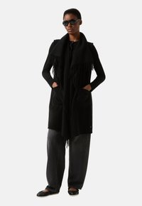 Sort tørklæde med frynser, båret over en sort bluse med lange ærmer. Løse grå jeans og sorte flade sko fuldender looket. Tilbehør inkluderer solbriller.