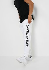 Białe spodnie dresowe z pionowym napisem „CALVIN KLEIN JEANS” w czerni na lewej nogawce, zestawione z czarnym T-shirtem i białymi trampkami.