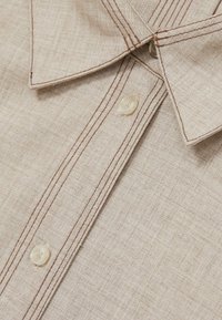 Beige knoopshirt van textiel stof, met bruine stiksels en doorschijnende plastic knopen op de sluiting en de kraag.