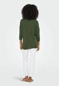 Top verde oliva, dal taglio morbido con maniche a tre quarti e orlo arrotondato, abbinato a pantaloni bianchi palazzo e sandali slip-on.