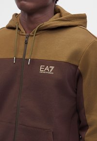 Άνδρας που φοράει καφέ και ελιά δίχρωμο φούτερ με φερμουάρ, με λογότυπο EA7 Emporio Armani στο στήθος, με ορατή κουκούλα με κορδόνι.