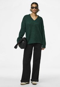 Zaļš, pārmērīgi liels adīts džemperis ar V veida kaklu, valkāts ar melniem platajiem biksēm un baltām kedām. Modelis tur melnu somu.