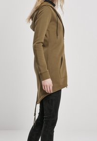 Olivově zelená dlouhá mikina s kapsou na přední straně, nastavitelnou šňůrkou a zakřiveným lemem. Kombinovaná s černými slim-fit kalhotami.