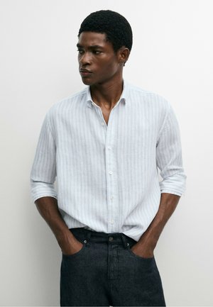 Giovane uomo con pelle scura e capelli corti e ricci, indossa una camicia bianca a righe a maniche lunghe e jeans scuri, mani in tasca, guarda a sinistra.