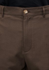 Κοντινή λήψη καφέ παντελονιών με ζώνη στη μέση με κουμπιά και ορατούς θηλιές για ζώνη, που δείχνει το μπροστινό μέρος στη λεκάνη.