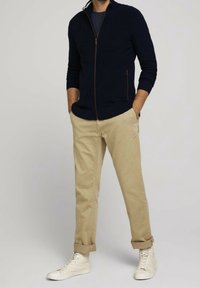Pull en tricot zippé bleu marine avec texture côtelée, chinos beiges et baskets montantes blanches, avec des manchons retroussés et des accents matériels minimalistes.