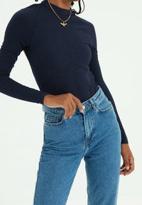 Haut bleu marine à manches longues côtelées avec un col montant et un motif en chevrons, associé à un jean taille haute bleu clair. Détail accentué par un collier en or.