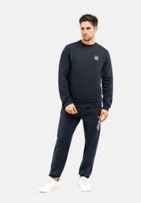 Felpa blu navy e pantaloni jogger abbinati in tessuto morbido, con un logo sul petto e testo grafico bianco sui pantaloni. Sneakers bianche.