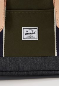 Grøn stofrygsæk med ferskenfarvede detaljer, der har et hvidt mærkelabel, blå denim-lignende tekstur i bunden og kontrastsyninger.