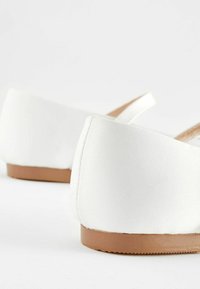Zapatillas de tela blanca con una textura suave, tacón bajo y suela de goma marrón. Presenta una forma redondeada con elementos de diseño minimalistas.