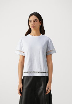 Mujer con largo cabello oscuro lleva una camisa blanca de manga corta con detalles de encaje en las mangas y el dobladillo, combinada con una falda de cuero negro.