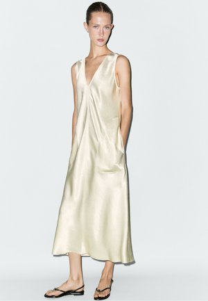 Donna che indossa un abito midi beige lucido senza maniche con tasche e sandali neri bassi, in piedi contro uno sfondo chiaro uniforme.