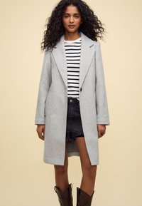 Manteau gris en laine, de coupe longue, avec des revers à cran et deux poches latérales, porté sur une chemise rayée et un short en denim noir, associé à des bottes marron.