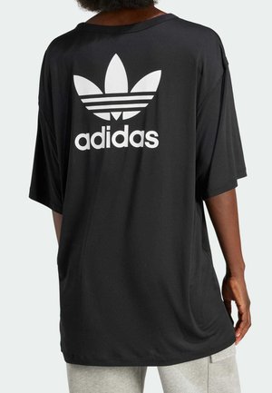 Person iført en oversize sort T-shirt med stort hvidt Adidas trefoil-logo og tekst på bagsiden, kombineret med lysegrå bukser.