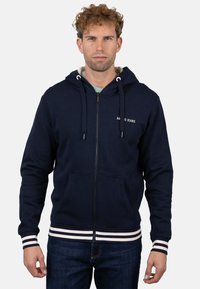 Sudadera con capucha azul marino con cremallera, capucha con cordón, dos bolsillos delanteros y puños y dobladillo acanalados con rayas blancas. Cuenta con el logo "AMMO JEANS" en el pecho.