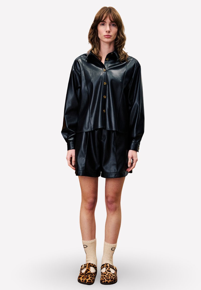 Zwarte faux leren overhemd met knopen, gecombineerd met bijpassende shorts. Slippersokken hebben een luipaardprint. Minimalistische, relaxte silhouette.