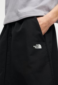 Χέρι στην τσέπη μαύρου σορτς The North Face που φοριέται με γκρι μπλουζάκι ορατό στη μέση.