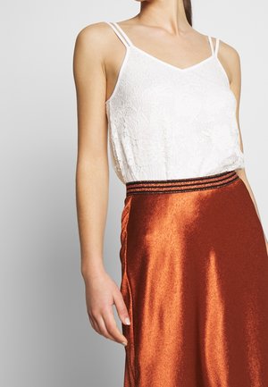 Kvinde iført hvid blondeærmeløs top og skinnende brændt orange nederdel med stribet taljebånd, arm og øvre torso synlige.