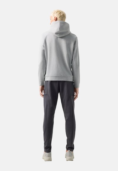 Personne aux cheveux blonds courts portant un sweat à capuche gris clair, un pantalon gris foncé et des baskets de couleur claire, debout de dos.