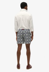 Mężczyzna ubrany w białą koszulę z długim rękawem oraz granatowo-białe szorty z kwiatowym wzorem, stoi tyłem na jednolitym tle.